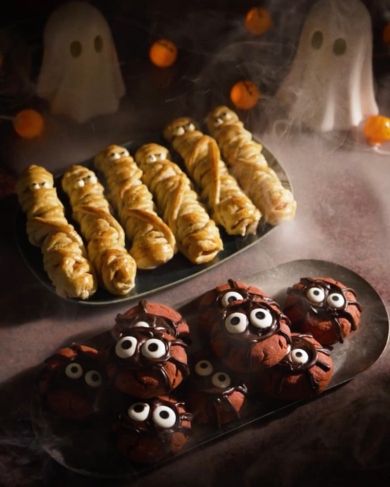 Momias de Frankfurt sin gluten y galletas araña Red Velvet en dos bandejas, con fantasmas y farolillos de calabaza de Halloween al fondo